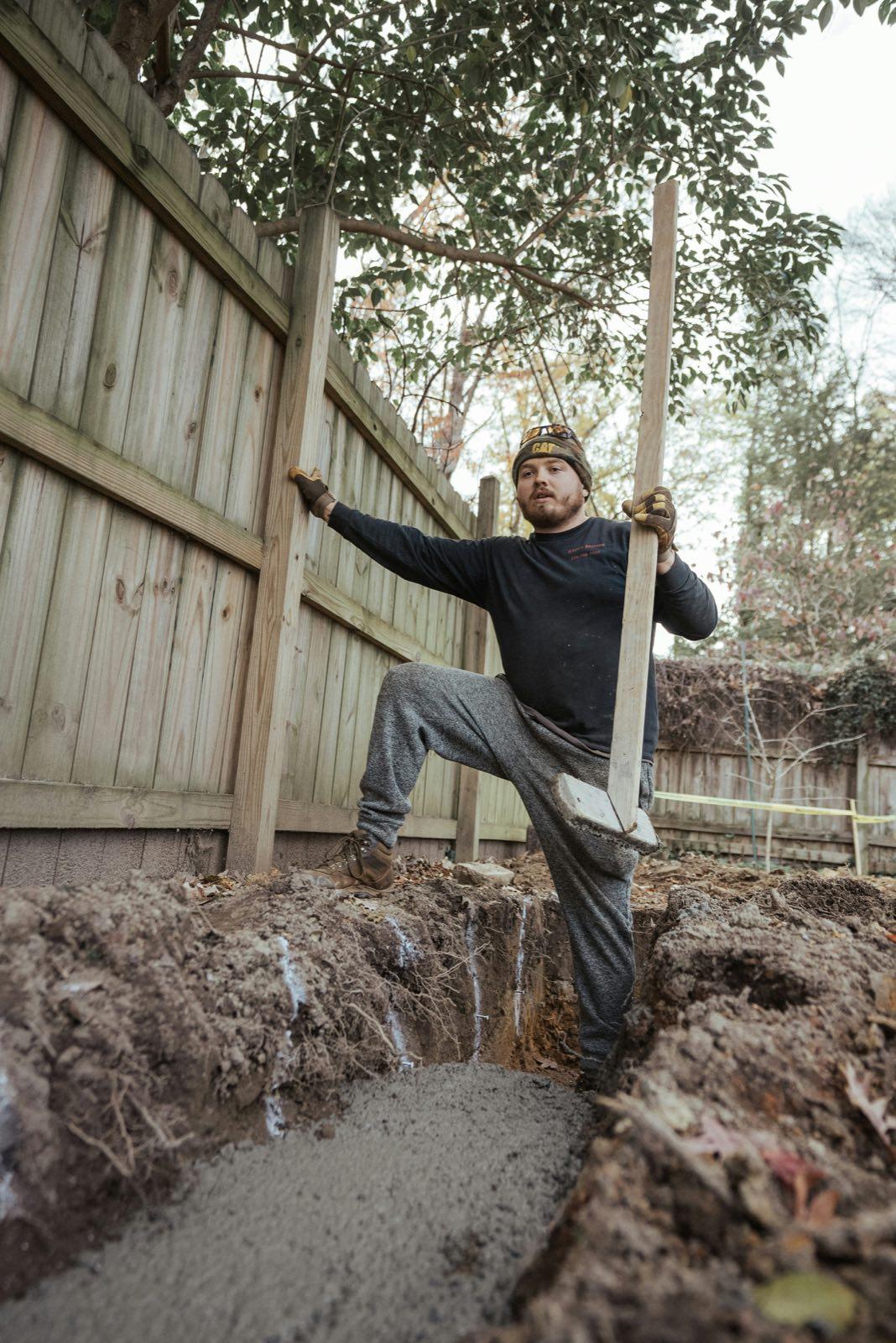 Worker holding a hand tamper over a freshly filled trench beside a wooden privacy fence