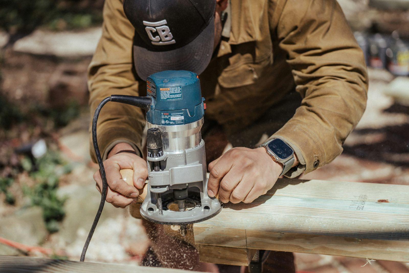 Close-up of carpenter in tan jacket and black cap running a router along a lumber board