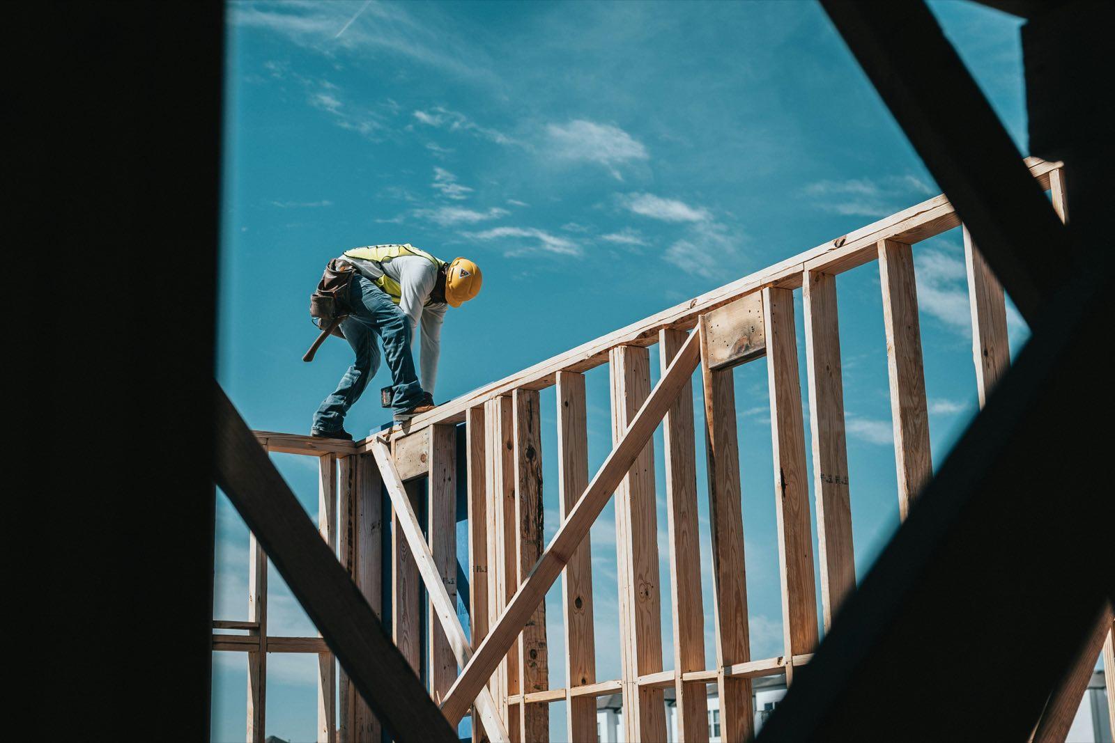 Framer in hard hat and safety vest standing atop a wood stud wall against blue sky