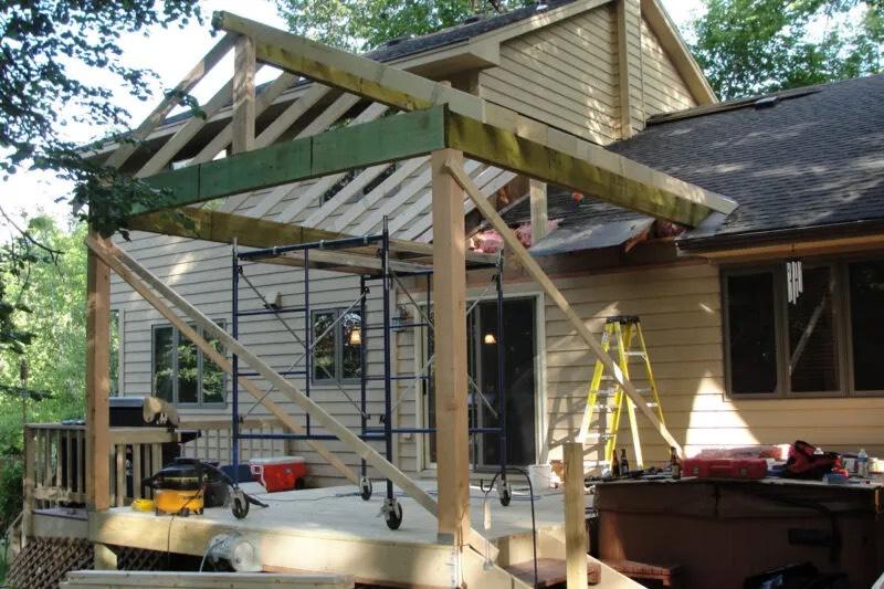 Porch roof framing in progress over a raised deck, with scaffolding and a yellow ladder