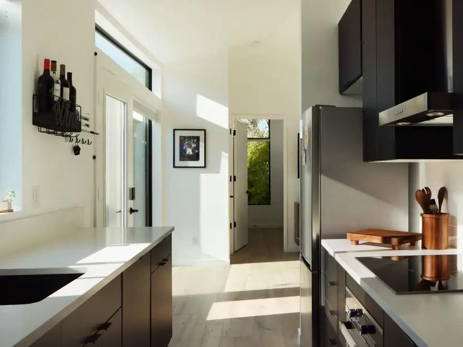 Galley kitchen with dark flat-panel cabinets, white counters, and stainless refrigerator beside a sunlit hallway