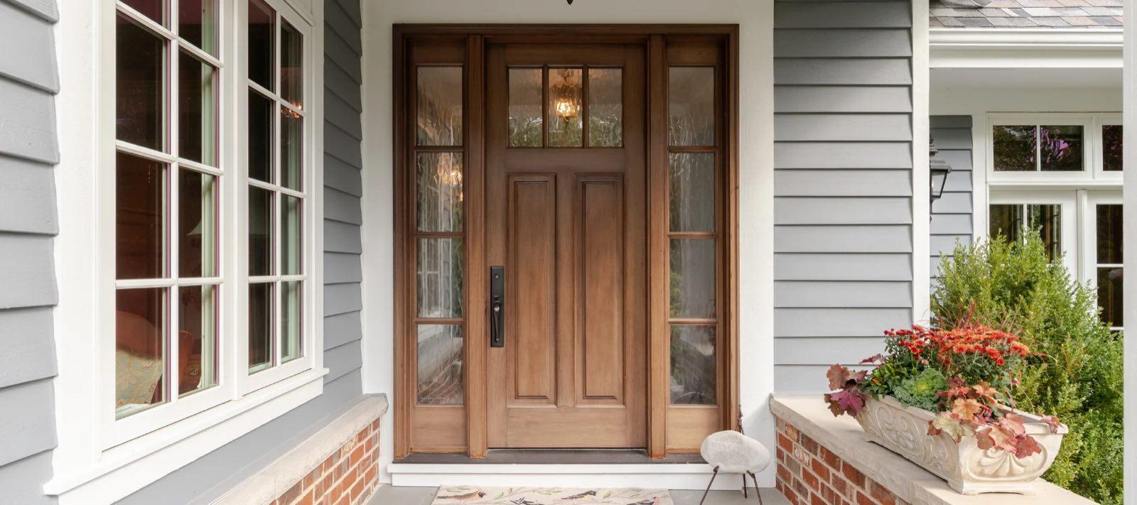 Stained-wood entry door with textured-glass sidelights on a gray clapboard house with brick planters