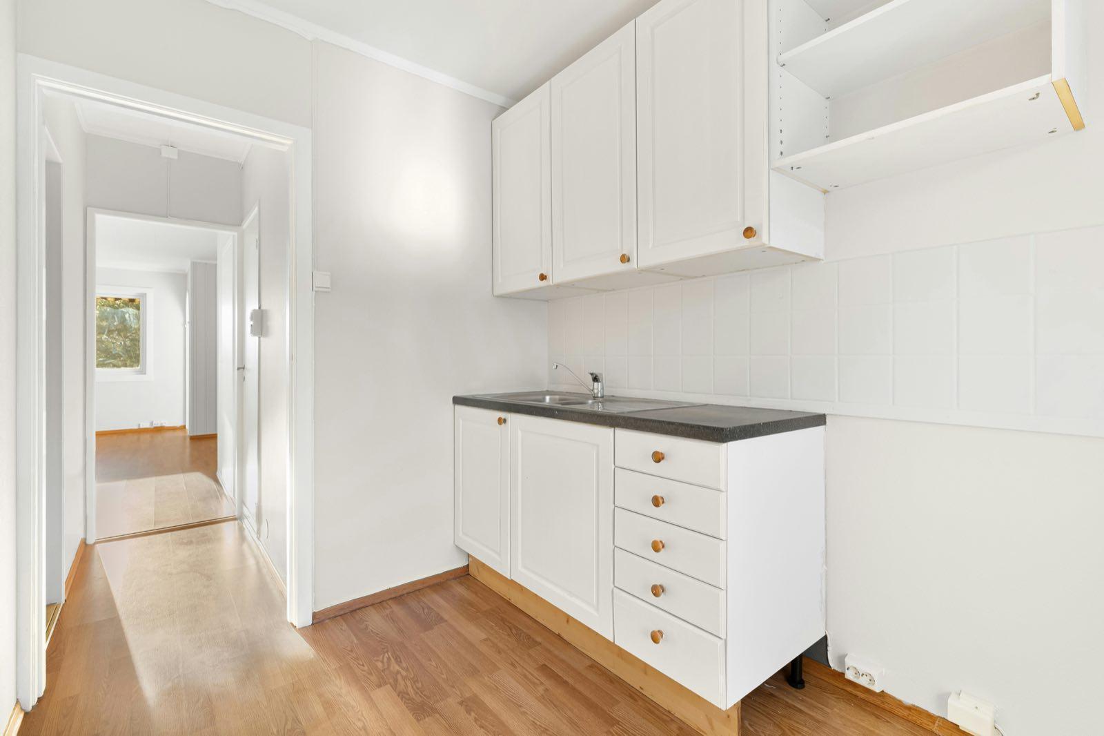 Small kitchenette with white cabinets and dark countertop on oak-look floor leading to a hallway