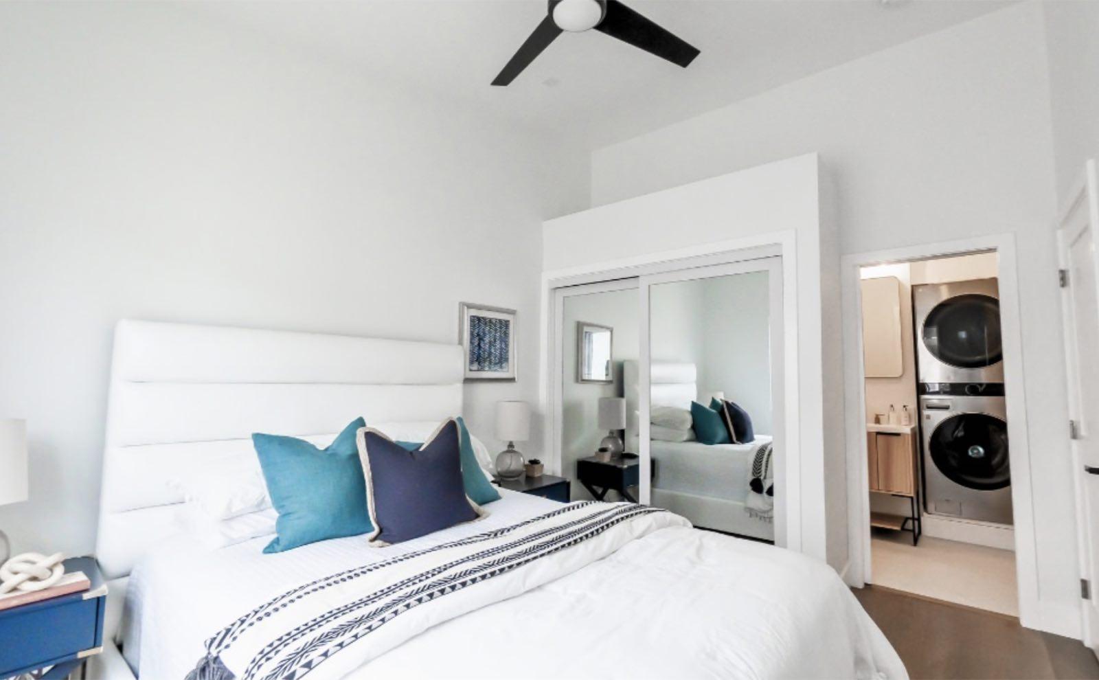 Bedroom with white upholstered headboard, mirrored closet doors, and stacked washer and dryer through doorway