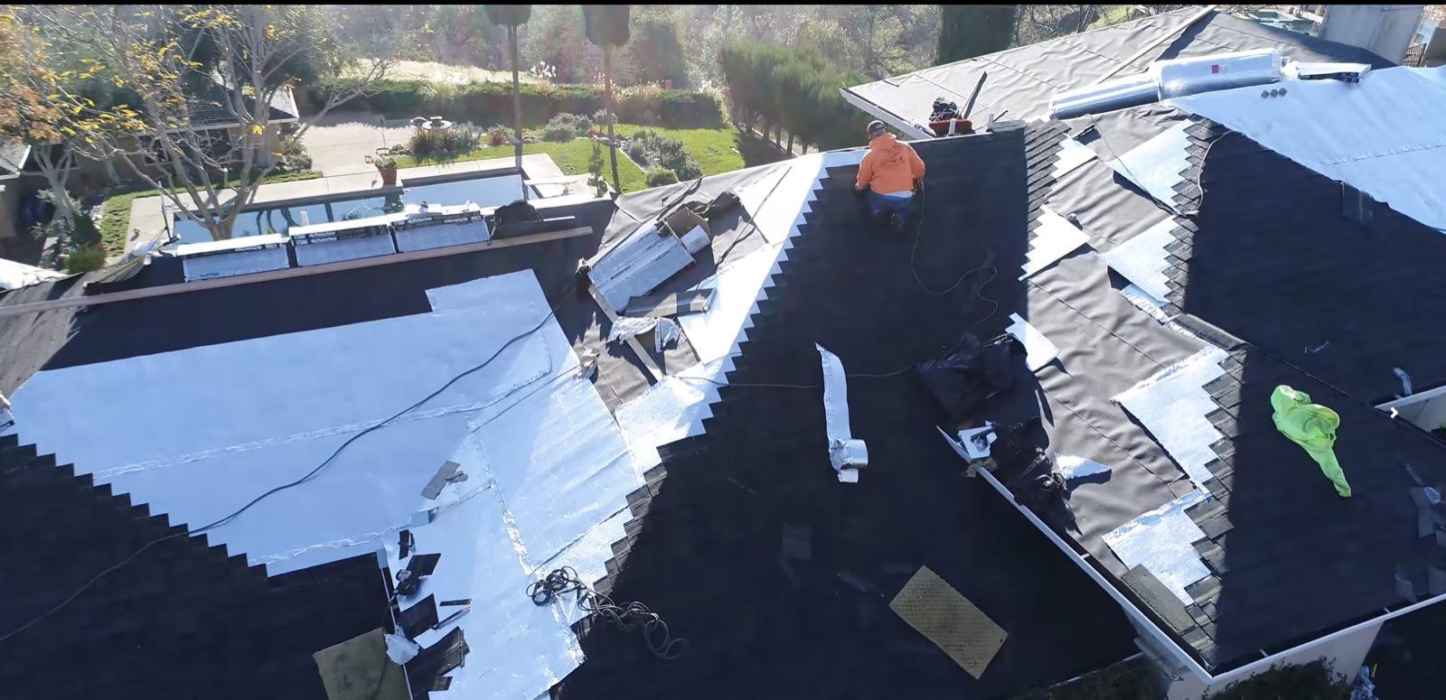 Aerial view of roofer in orange shirt laying dark shingles over silver underlayment