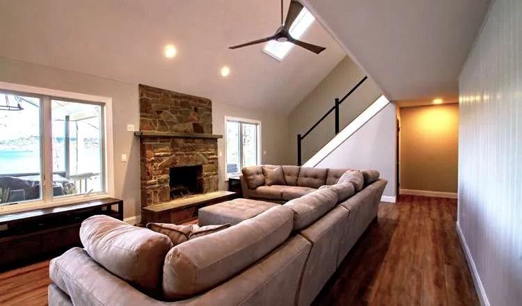 Taupe sectional facing a rustic stone fireplace beneath a vaulted ceiling with skylight and fan