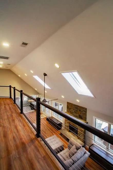 View from loft with cable railing over vaulted living room with skylights and stone fireplace
