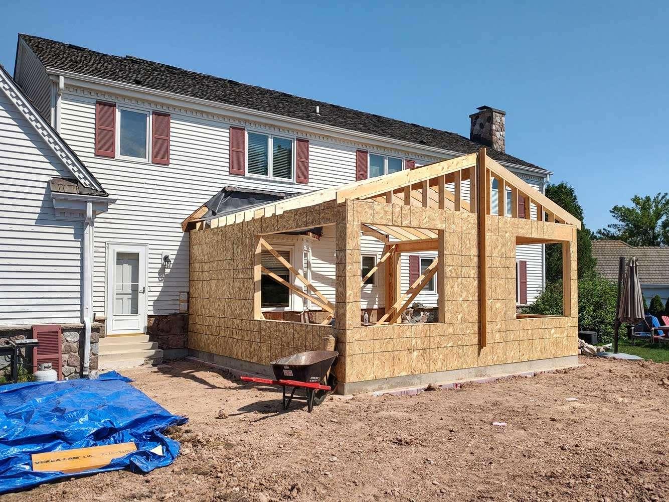 Single-story addition under construction with OSB sheathing and open roof framing on a white house