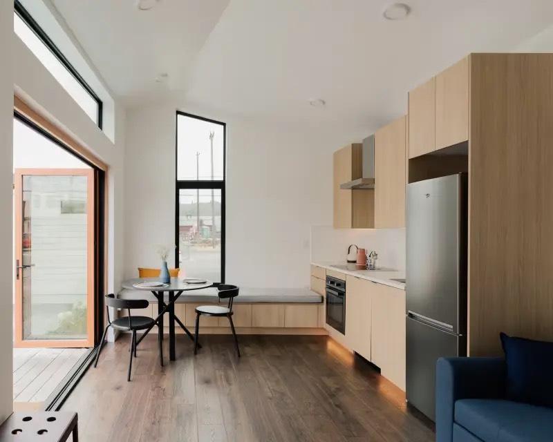 Compact studio with light oak kitchenette, stainless refrigerator, built-in bench, and tall black-framed window