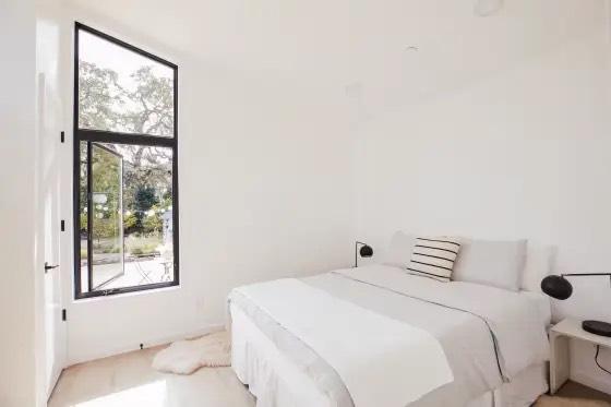 Bright white bedroom with gray bedding, black wall sconces, and tall black-framed window