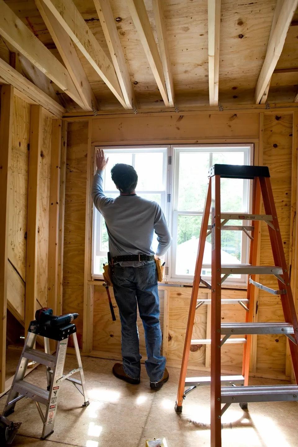 Carpenter in tool belt checking a newly set window in an unfinished plywood-sheathed room