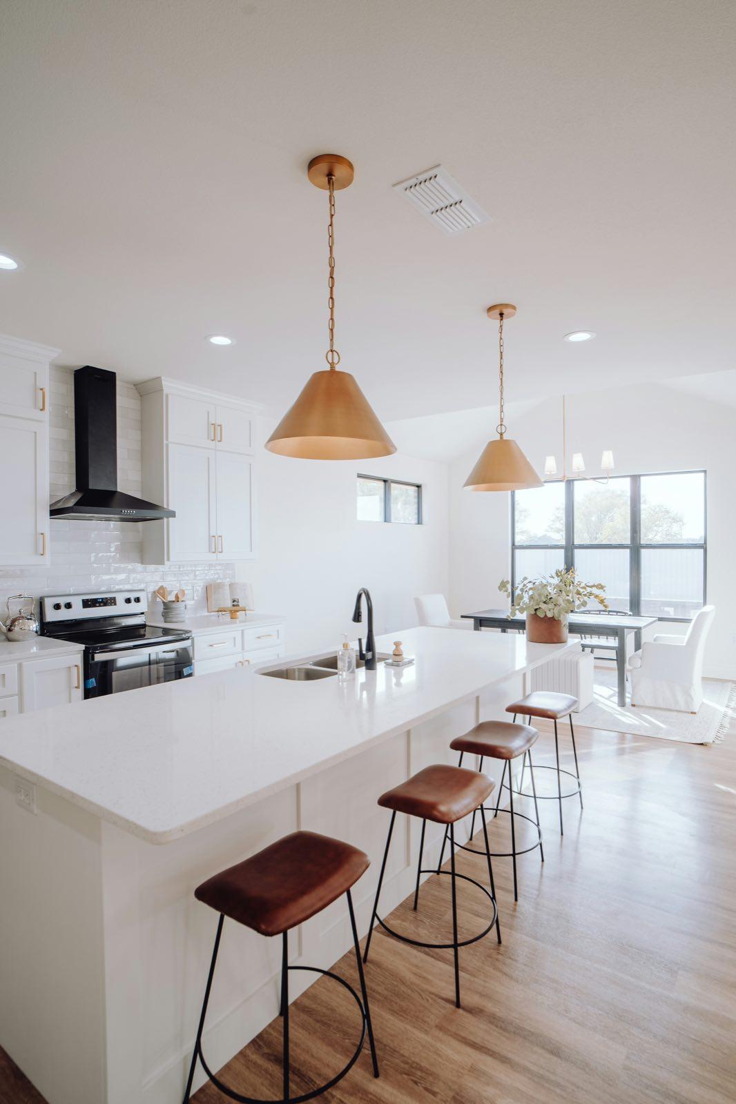 White kitchen with long white-topped island, leather bar stools, brass cone pendants, and black range hood