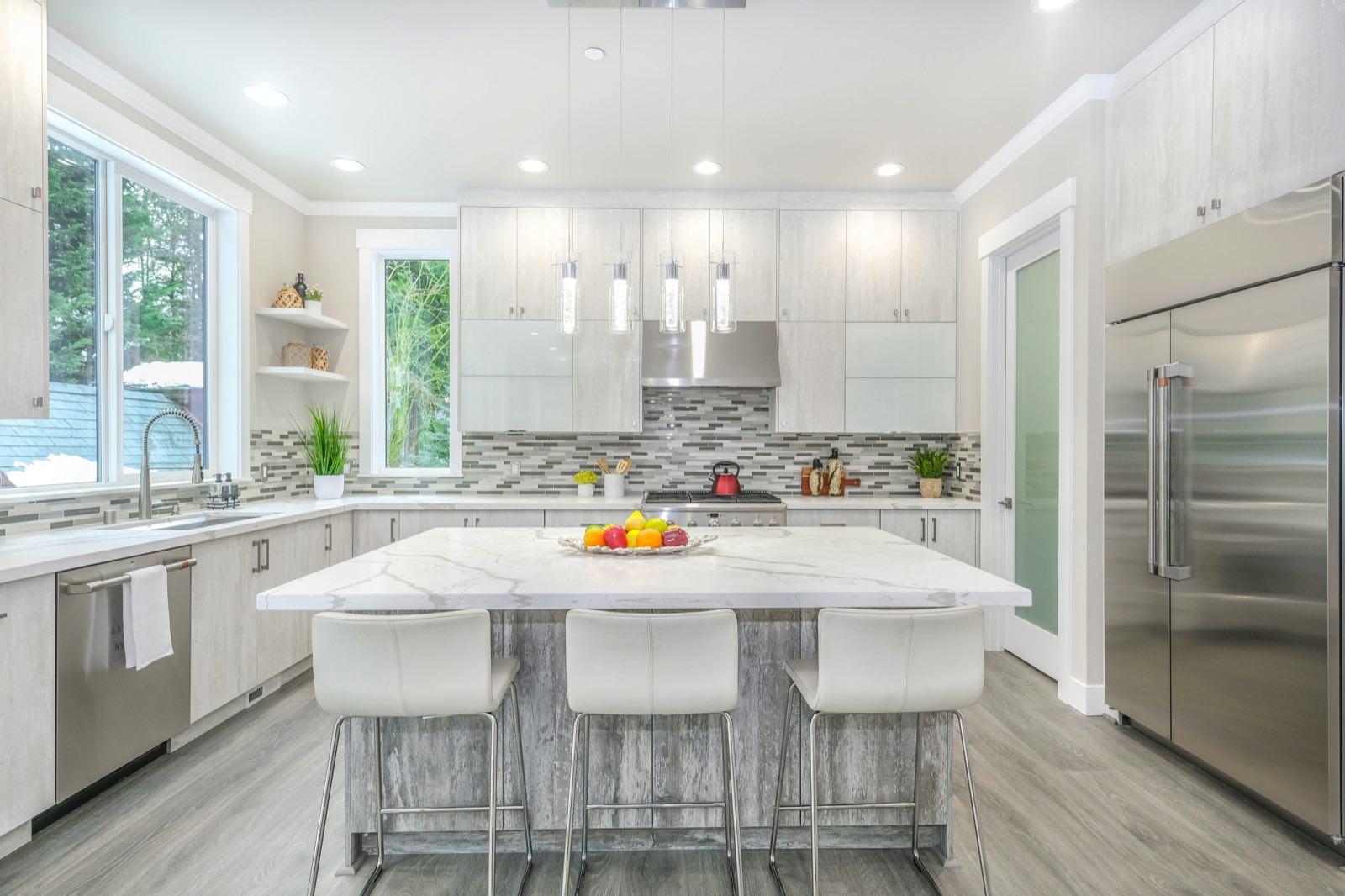 Kitchen island with marble-look top and white stools beneath glass pendants, facing mosaic tile backsplash